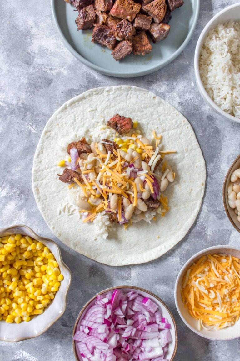 Open steak burrito in the process of being built surrounded by bowls of fillings.