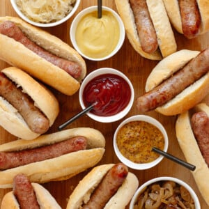 Wooden board covered with grilled brats in buns and small bowls of various toppings.