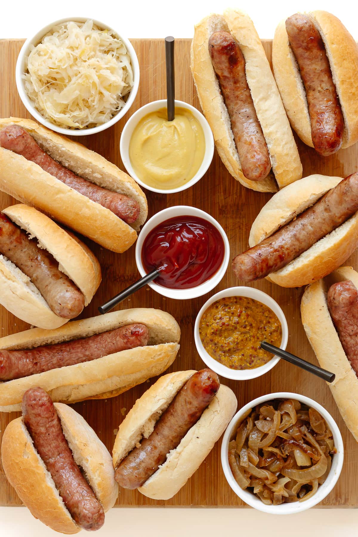 Wooden board covered with grilled brats in buns and small bowls of various toppings.