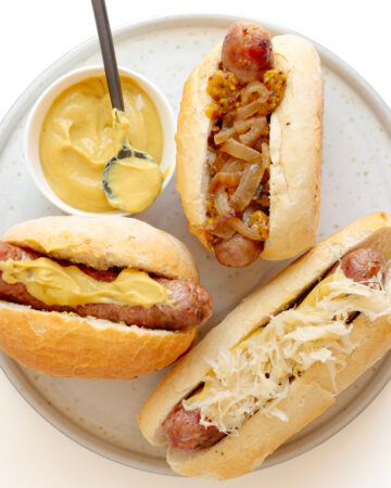 Three grilled bratwurst on buns with different toppings sitting on a plate with a small bowl of German mustard.
