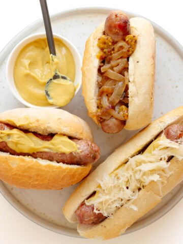 Three grilled bratwurst on buns with different toppings sitting on a plate with a small bowl of German mustard.