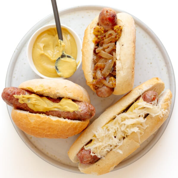 Three grilled bratwurst on buns with different toppings sitting on a plate with a small bowl of German mustard.