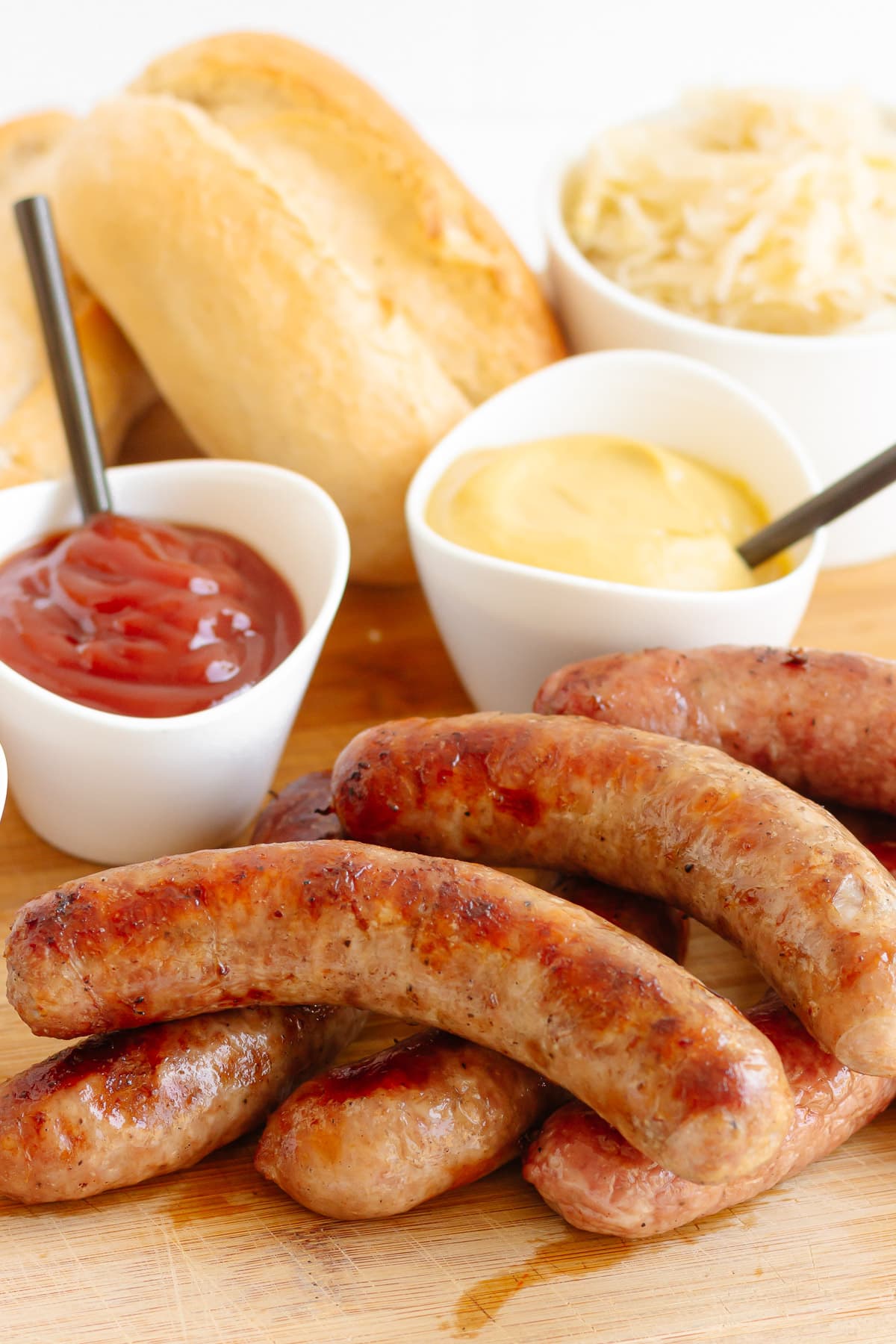 Grilled bratwurst on a wooden board with a pile of crusty buns and small bowls of toppings.