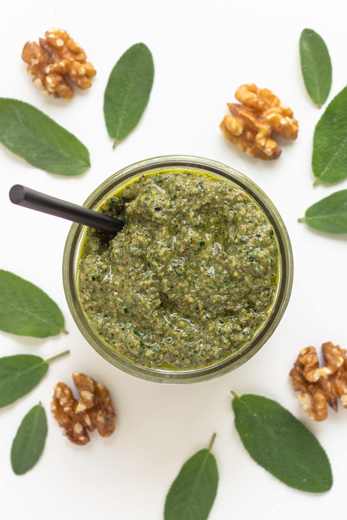 Small bowl of sage pesto surrounded by fresh sage leaves and walnut pieces.