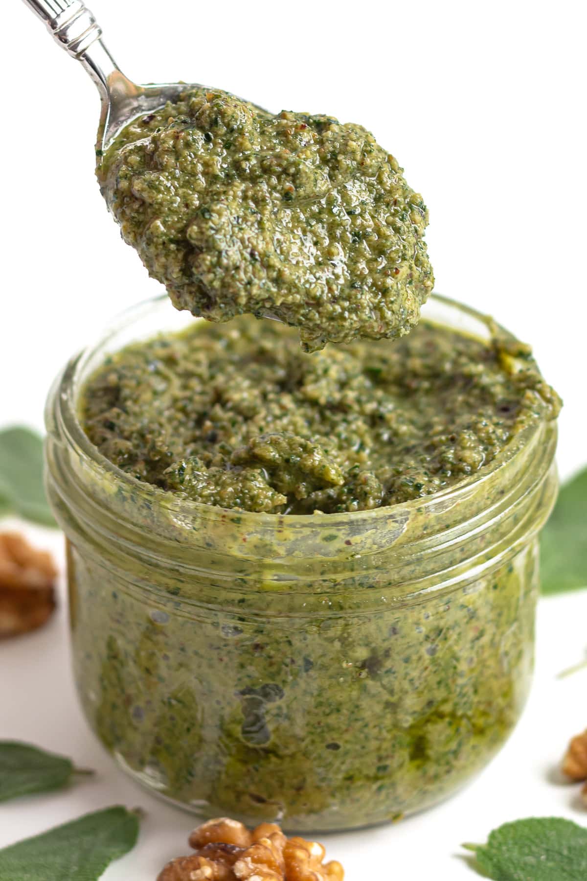 Spoonful of sage pesto lifting out of glass jar surrounded by sage leaves and walnuts.