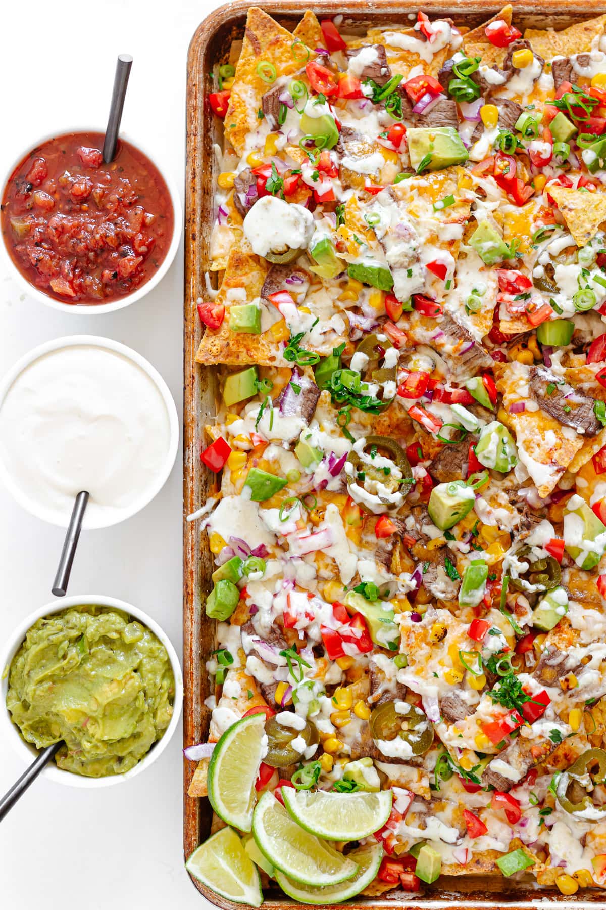 Steak nachos on a sheet pan with small bowls of salsa, sour cream and guacamole beside it.