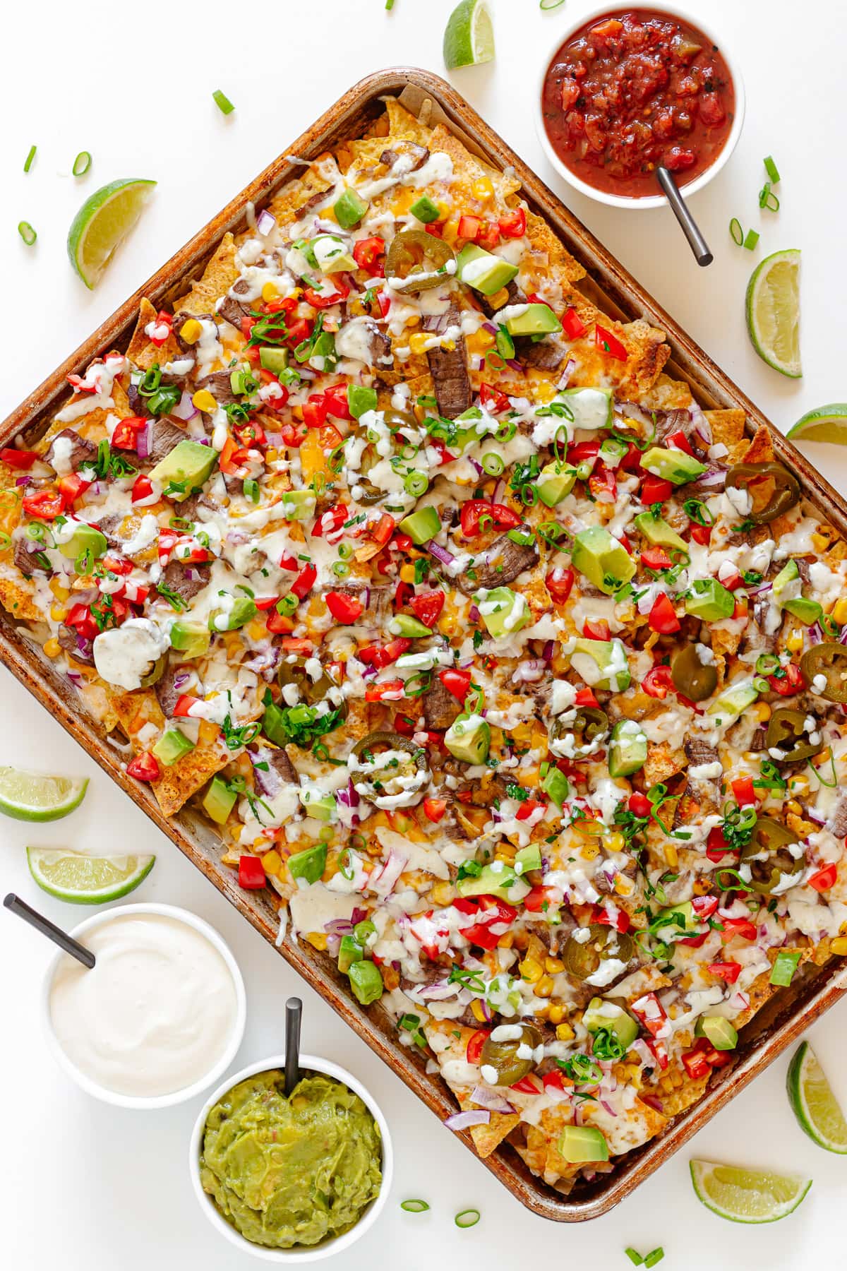 Sheet pan of steak nachos surrounded by lime wedges and small bowls of salsa, sour cream and guacamole.