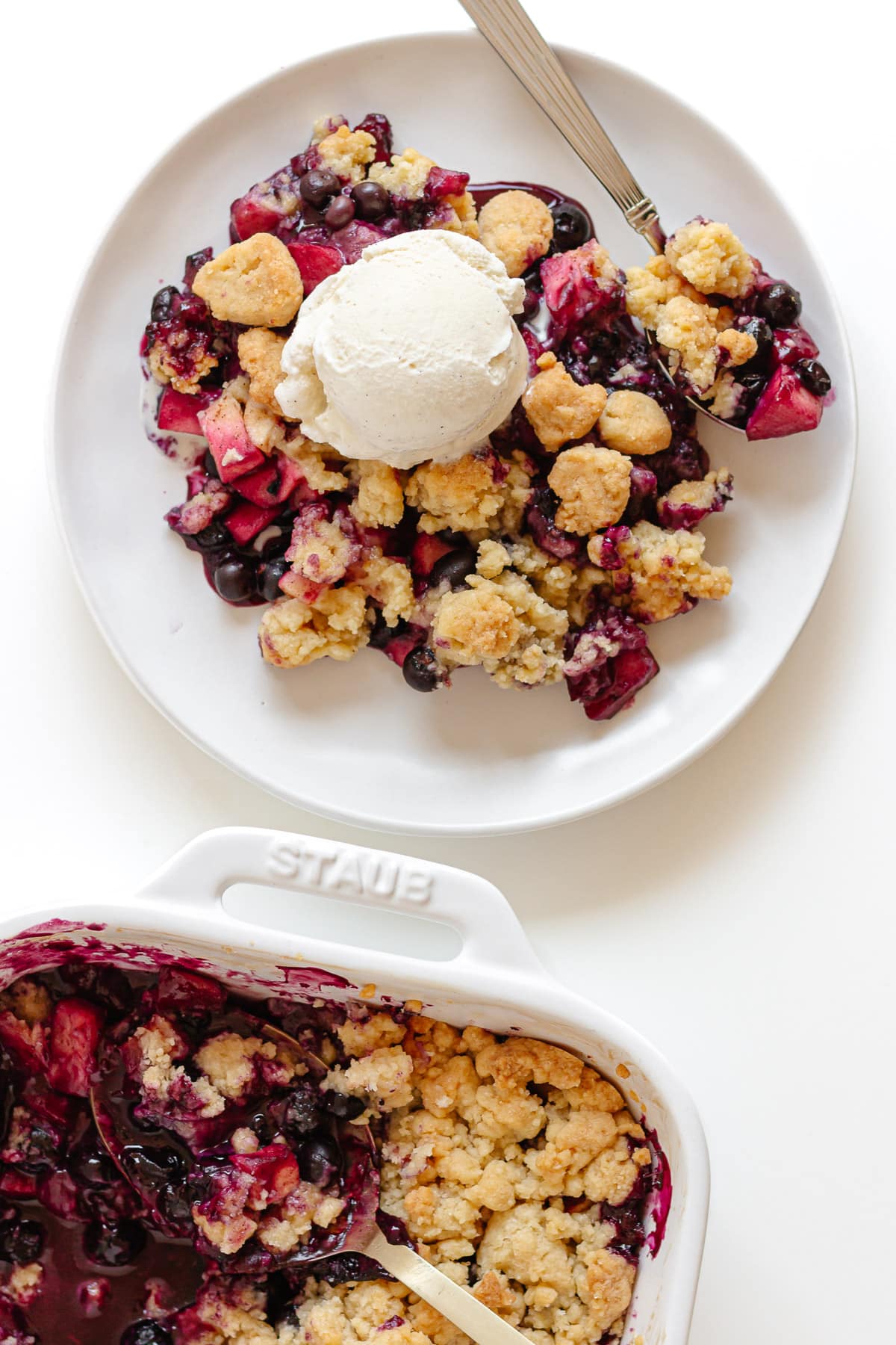 Plate of apple blueberry crumble topped with vanilla ice cream next to the baking dish it was scooped out from.