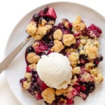 Blueberry and apple crumble on a white plate with spoon and a scoop of vanilla ice cream on top.