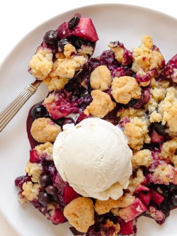 Blueberry and apple crumble on a white plate with spoon and a scoop of vanilla ice cream on top.