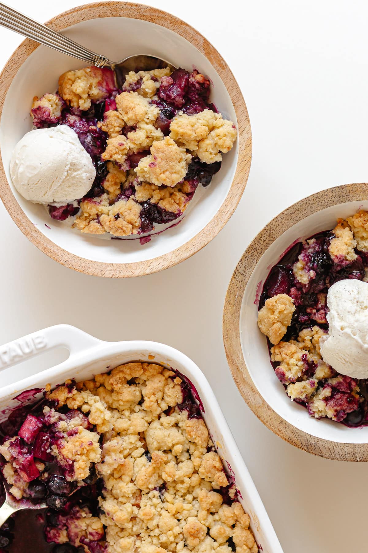 Two bowls of blueberry apple crumble topped with vanilla ice cream next to the baking dish.
