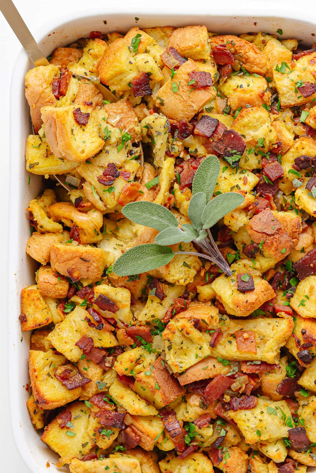 Casserole dish of stuffing with serving spoon and sprig of fresh sage.