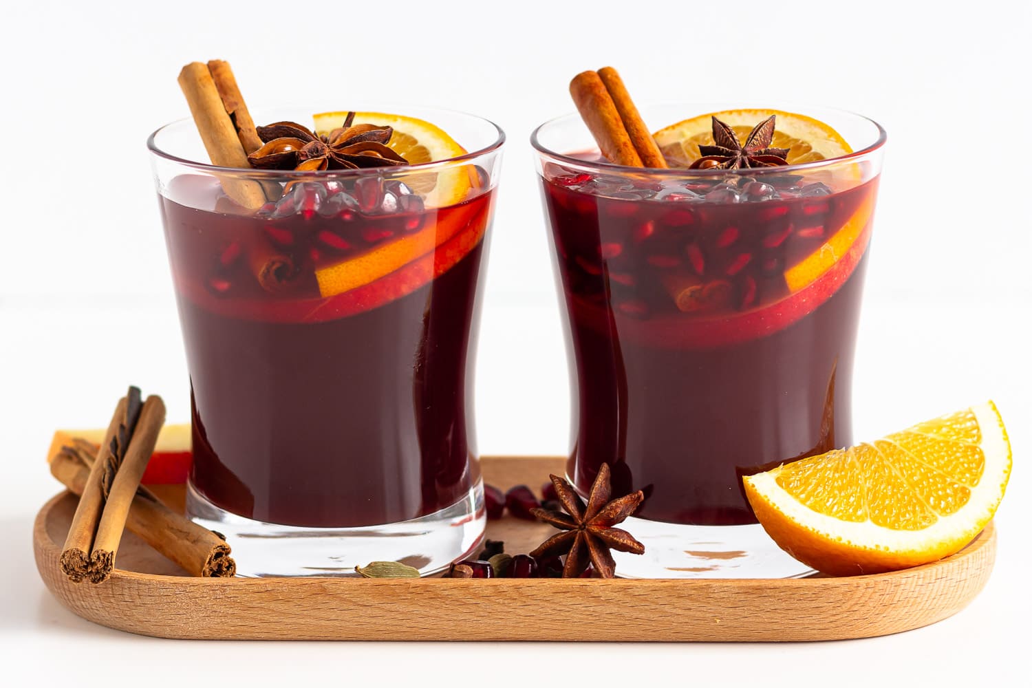 Two glasses of Kinderpunsch on a wooden tray garnished with orange slices, cinnamon sticks, star anise and pomegranate arils.