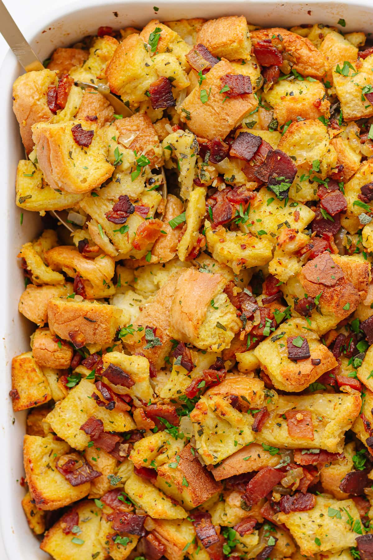 Serving spoon digging into a casserole dish of sage and onion stuffing with bacon.