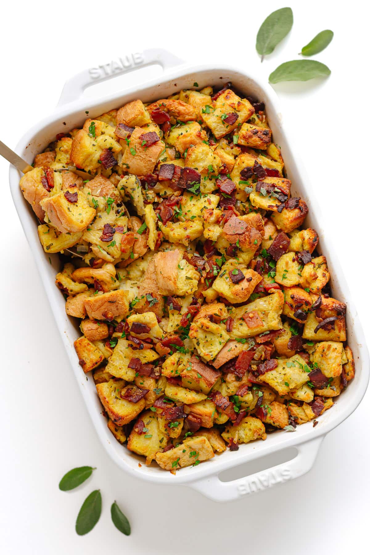 Stuffing in a white casserole dish with serving spoon and fresh sage leaves scattered next to it.