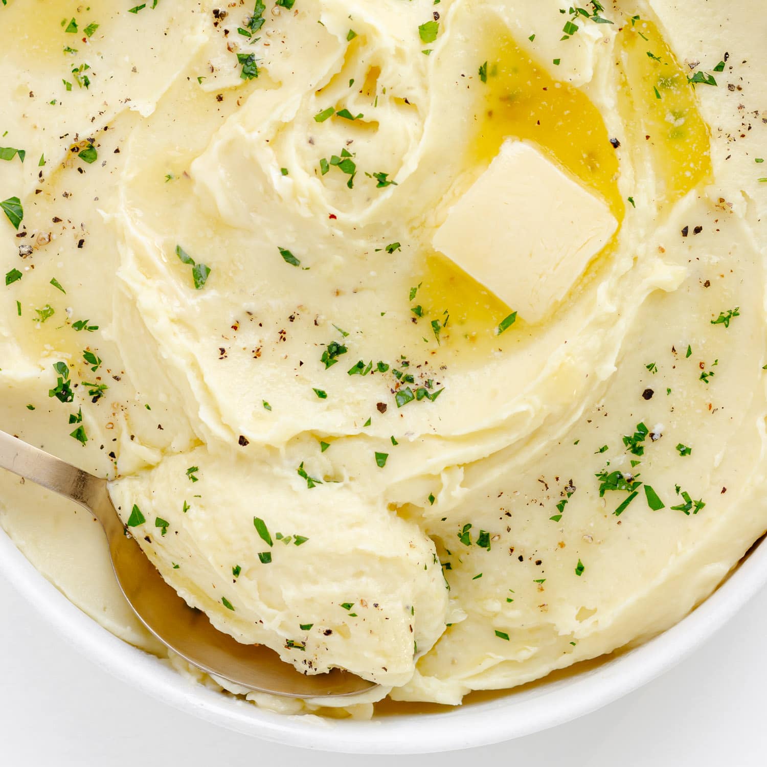 Serving spoon in a bowl of mashed potatoes garnished with melted butter, chopped parsley and cracked pepper.