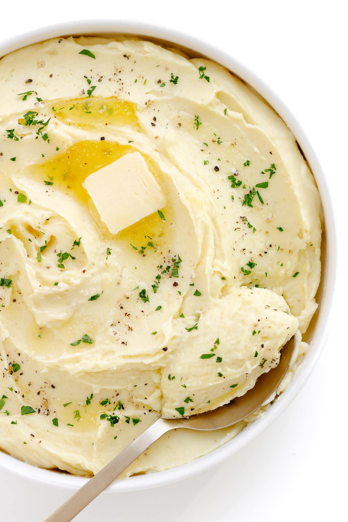 Whipped mashed potato in serving bowl with pat of butter, chopped parsley and black pepper for garnish.