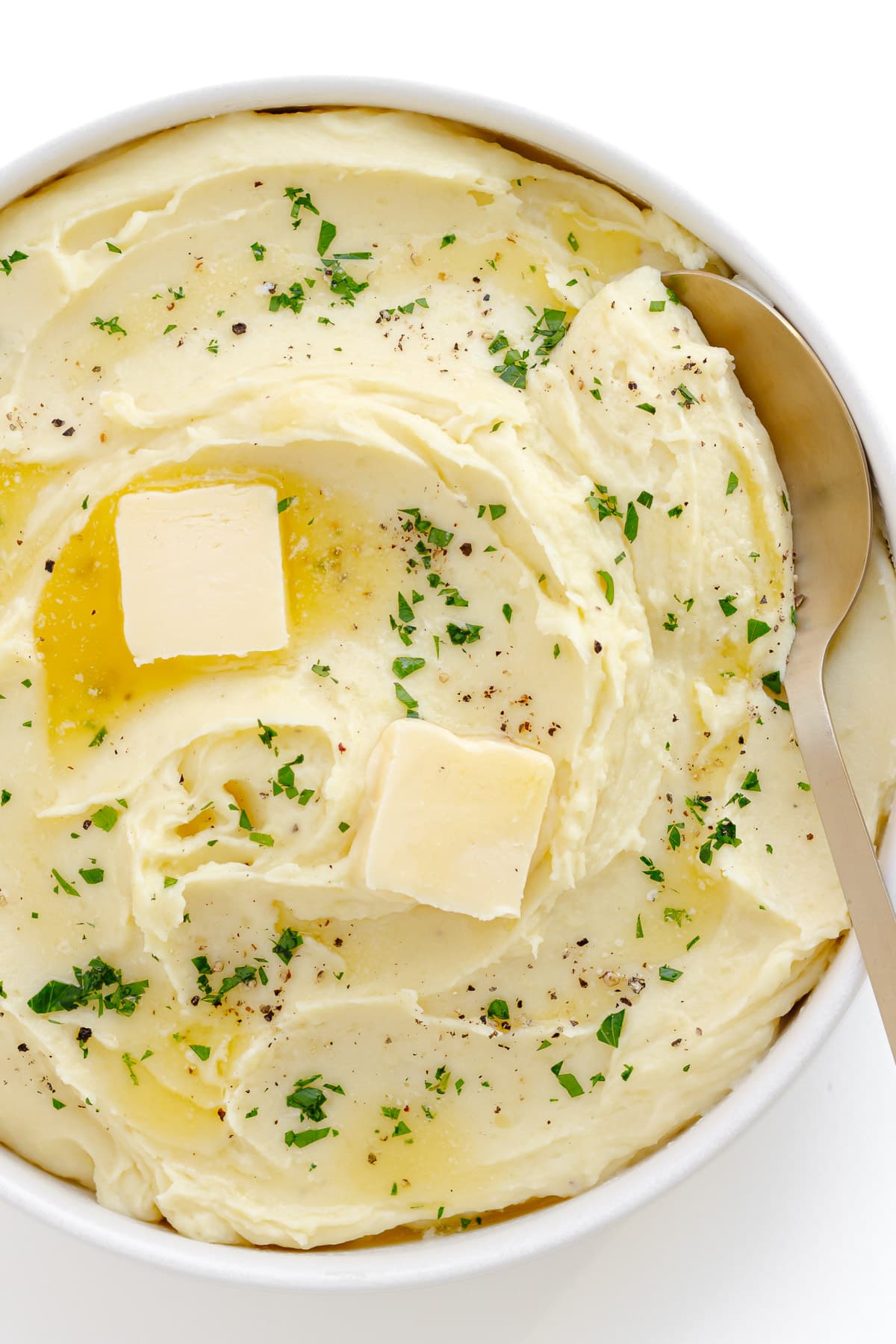 Whipped mashed potatoes in a white serving bowl garnished with pats of butter, chopped parsley and cracked black pepper.