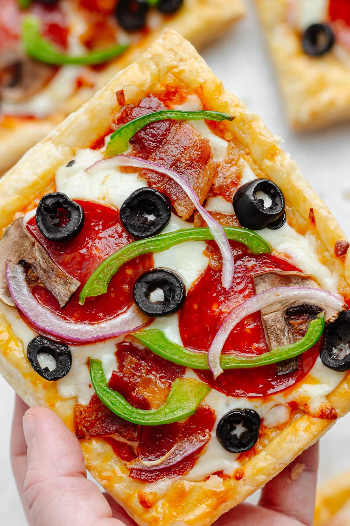 Hand holding a square puff pastry pizza topped with tomato sauce, mozzarella, pepperoni, bacon, green bell pepper, mushrooms, red onion and black olives.