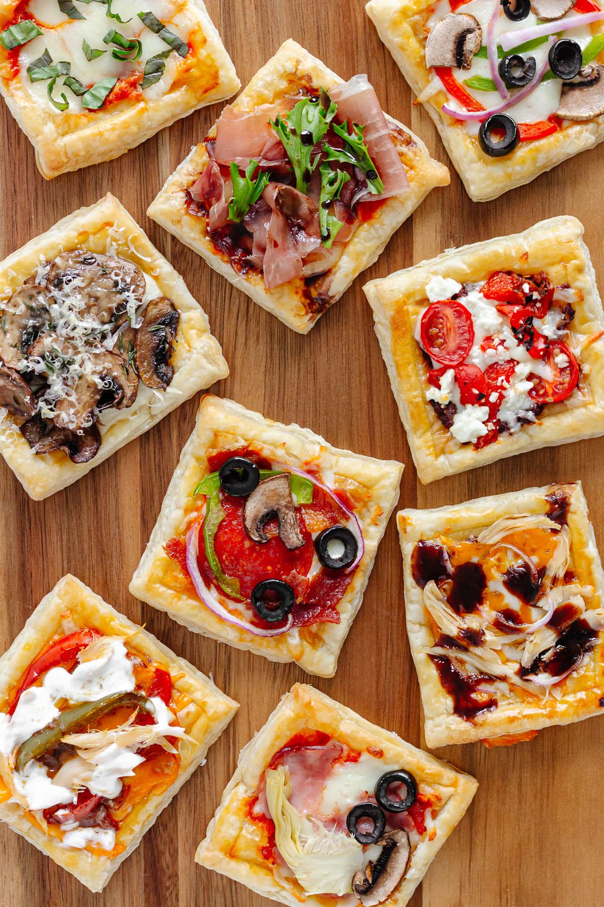 A variety of small puff pastry pizzas with different toppings on a wooden serving board.