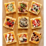 Nine small puff pastry pizzas with different toppings on a wood board.