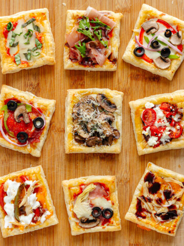 Nine small puff pastry pizzas with different toppings on a wood board.
