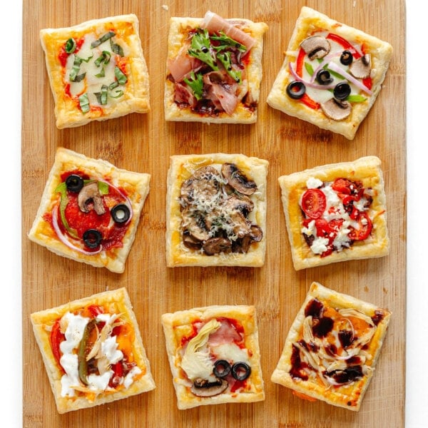 Nine small puff pastry pizzas with different toppings on a wood board.