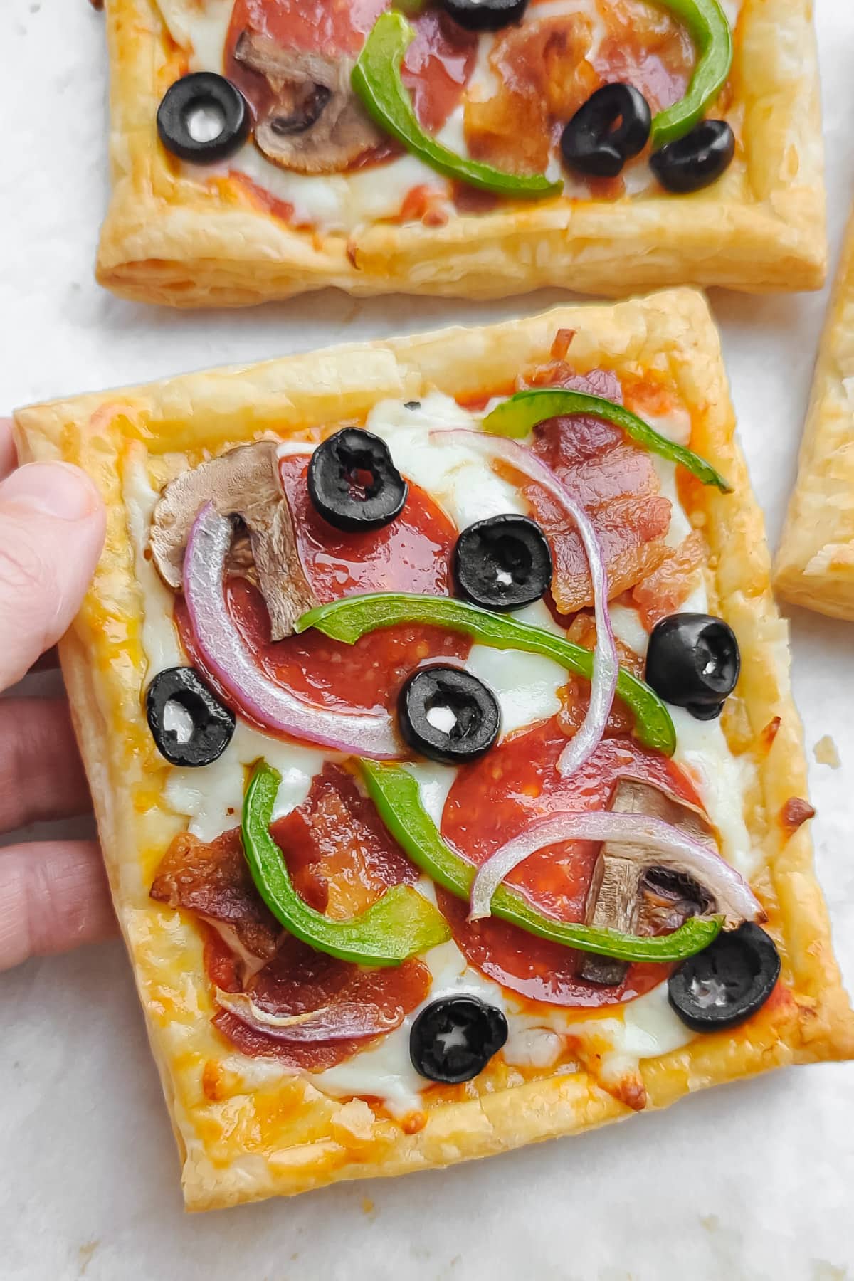 Hand lifting up a square slice of supreme puff pastry pizza.