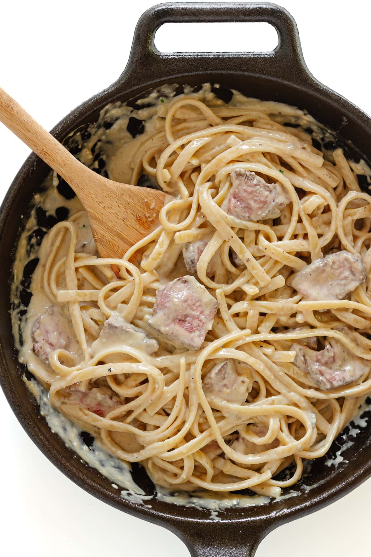 Creamy steak pasta in cast iron skillet with wooden spoon.