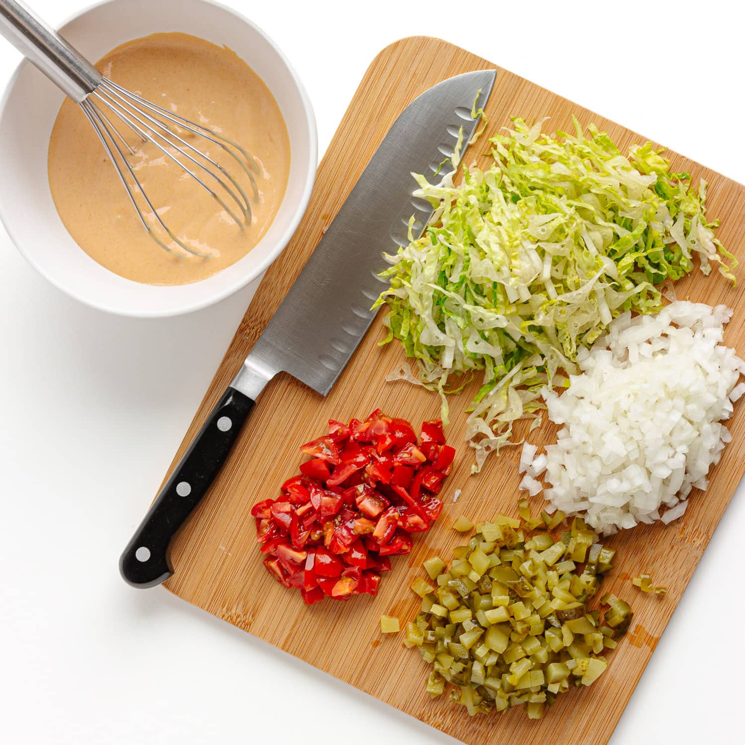 Homemade burger sauce in white bowl with whisk next to cutting board with prepped burger toppings (shredded lettuce, chopped onions, pickles and tomatoes).