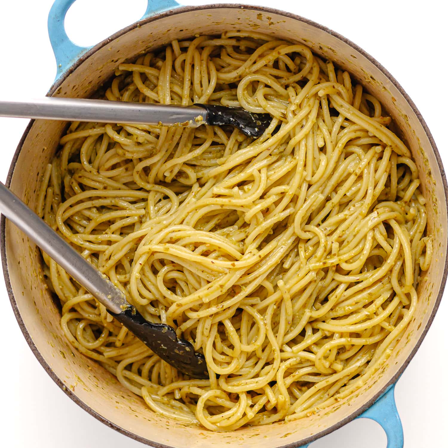 Tongs tossing spaghetti with pesto in a dutch oven.