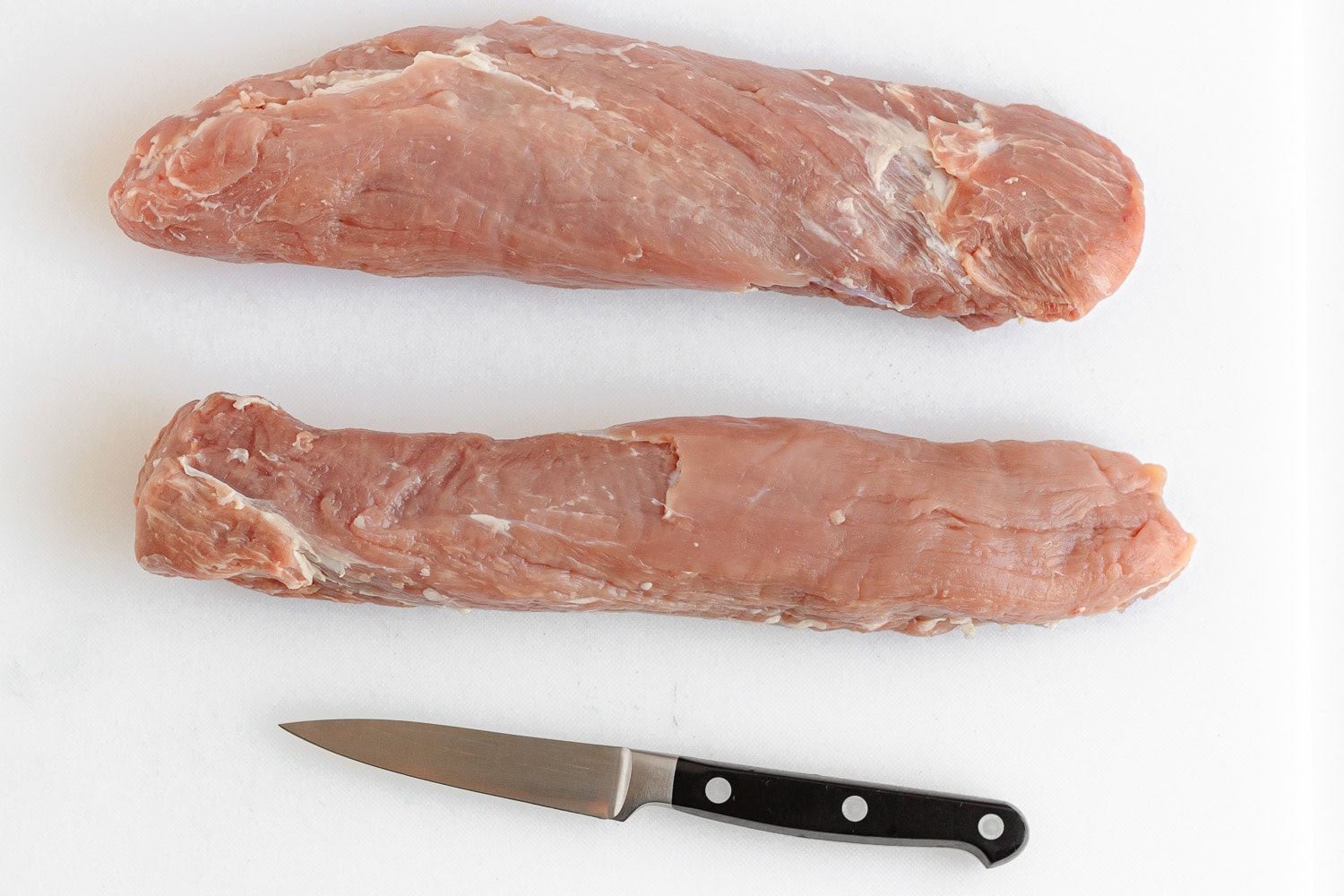 Two trimmed pork tenderloin on a white plastic cutting board with knife.
