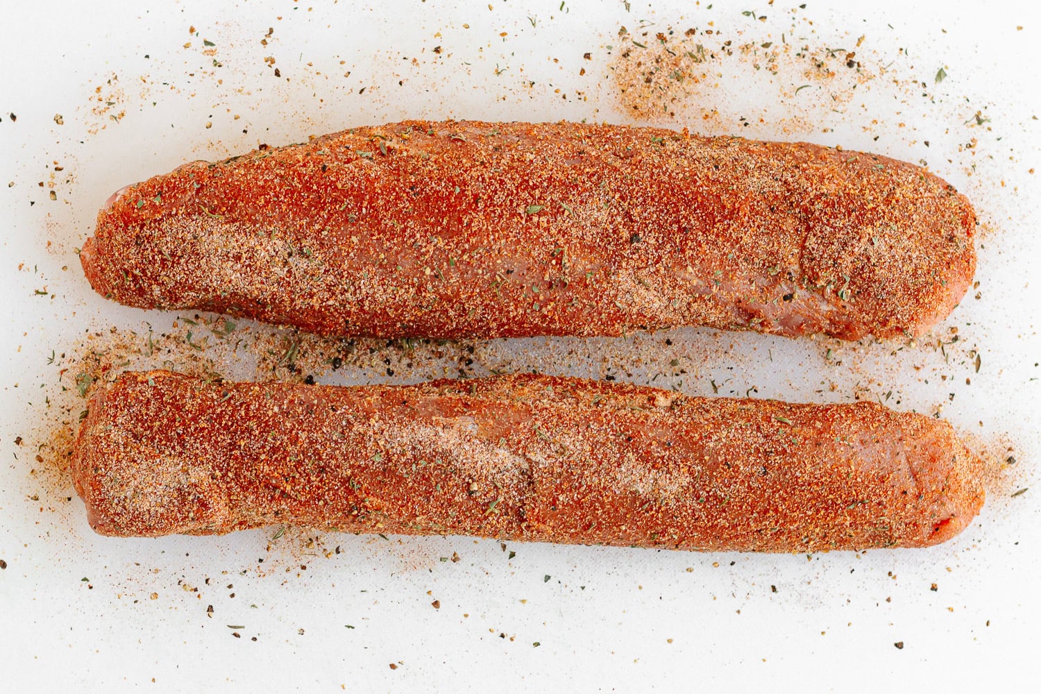 Two seasoned pork tenderloin on a white cutting board.