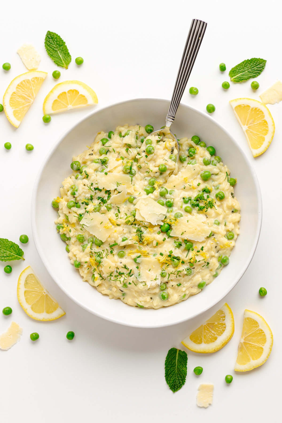 White bowl of pea risotto on a white surface with lemon slices, peas, mint and parmesan shavings scattered around.