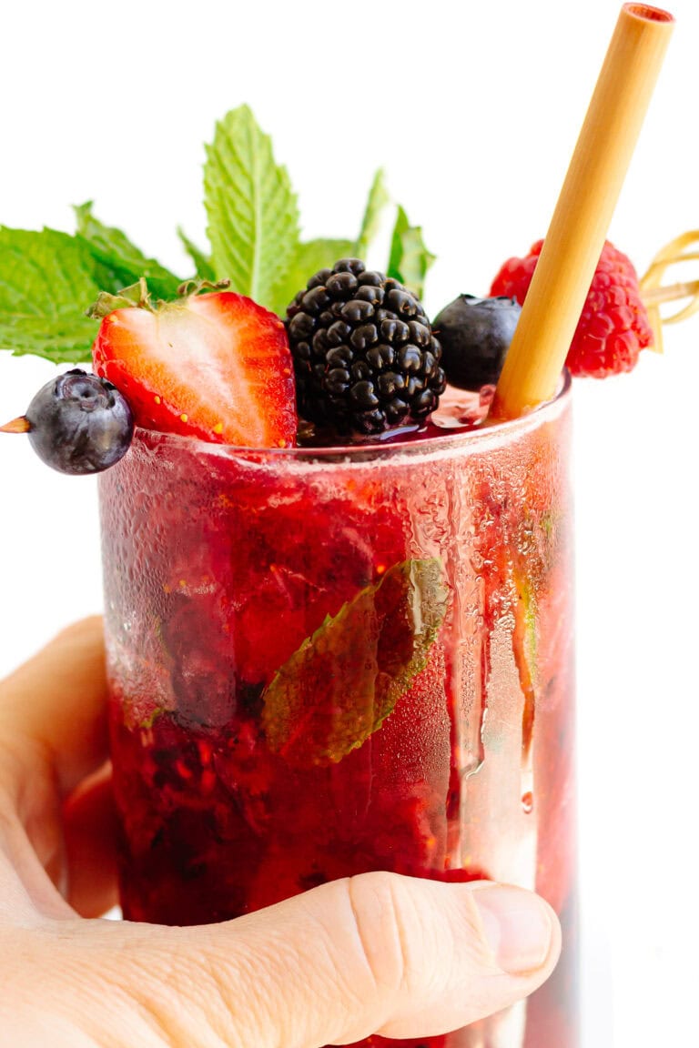 Hand holding a berry mojito cocktail garnished with fresh mint and mixed berries on a cocktail skewer.