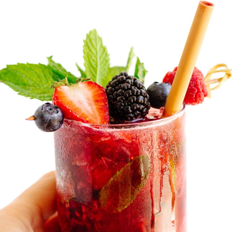 Hand holding a berry mojito garnished with fresh mint, mixed berries on a cocktail skewer and a bamboo straw.