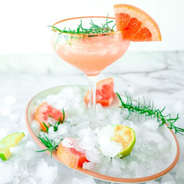 A pink grapefruit rosemary cocktail on a tray covered with ice, grapefruit, lime and rosemary.