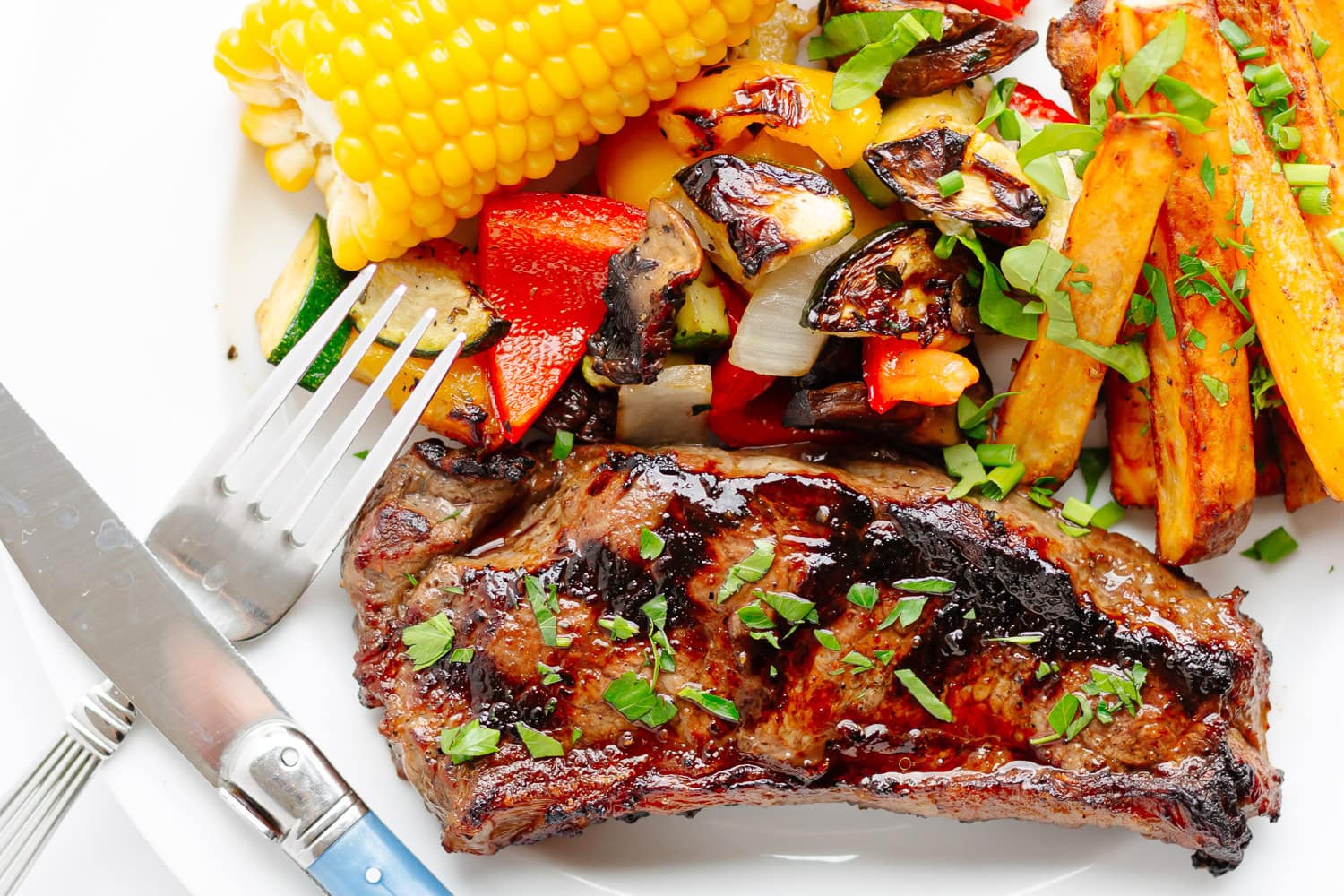 Grilled steak on a plate along with a few sides (corn, mixed grilled veggies and potato wedges).