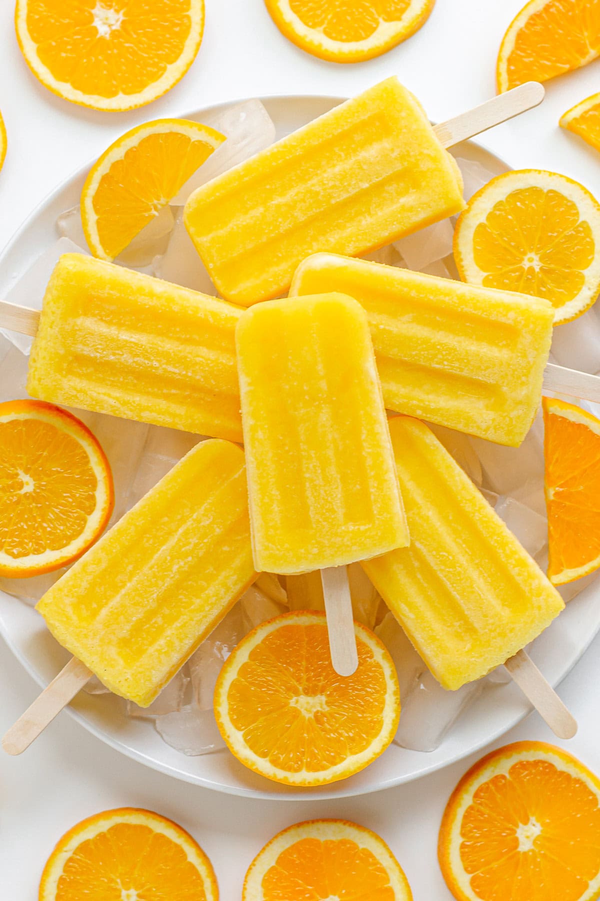 Orange popsicles piled on a plate of ice surrounded by orange slices.