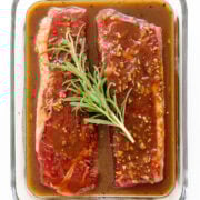 Two steaks in a glass dish covered in steak marinade with a sprig of fresh rosemary on top.