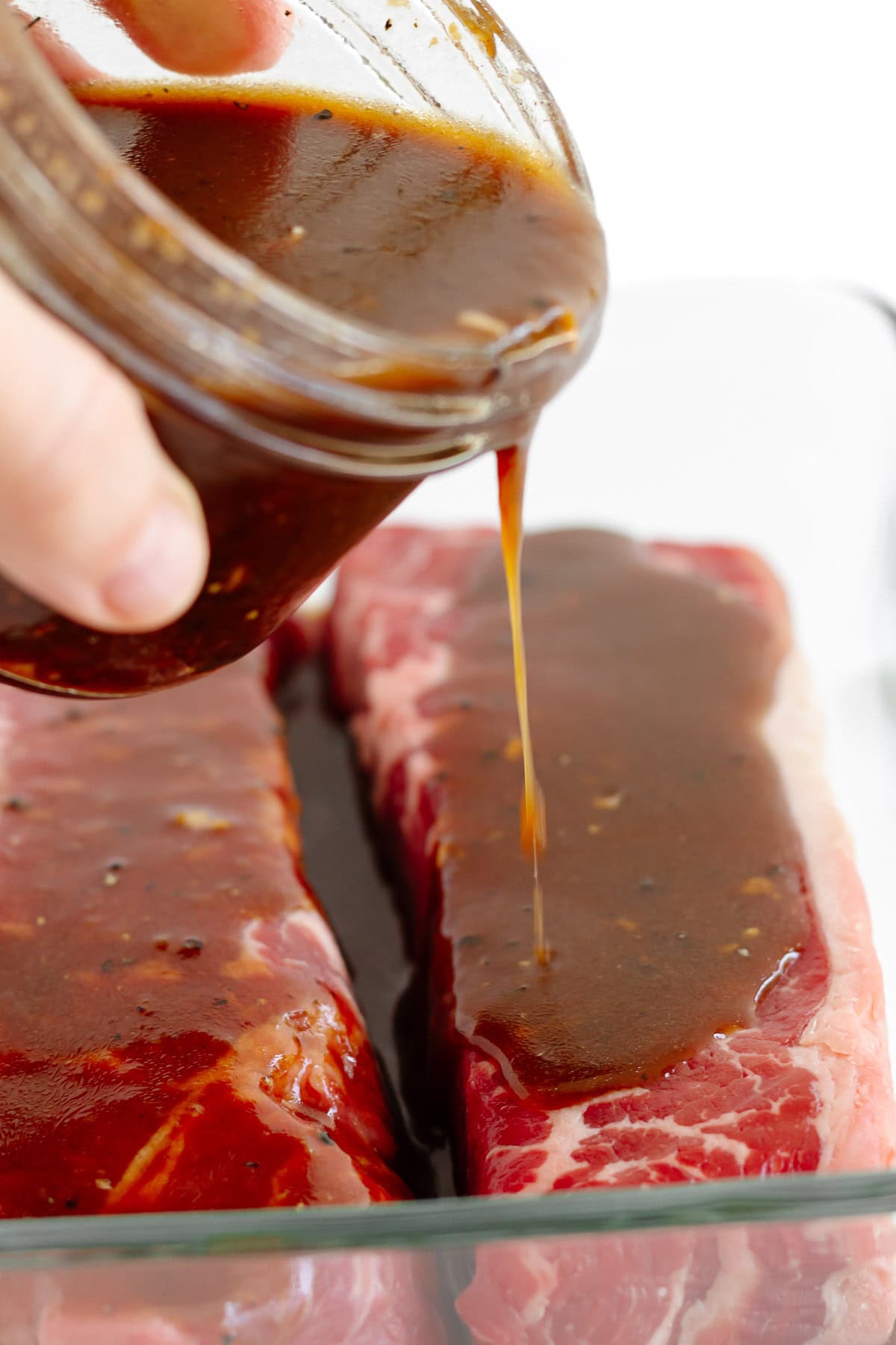 Hand pouring small jar of steak marinade over two steaks in a glass dish.