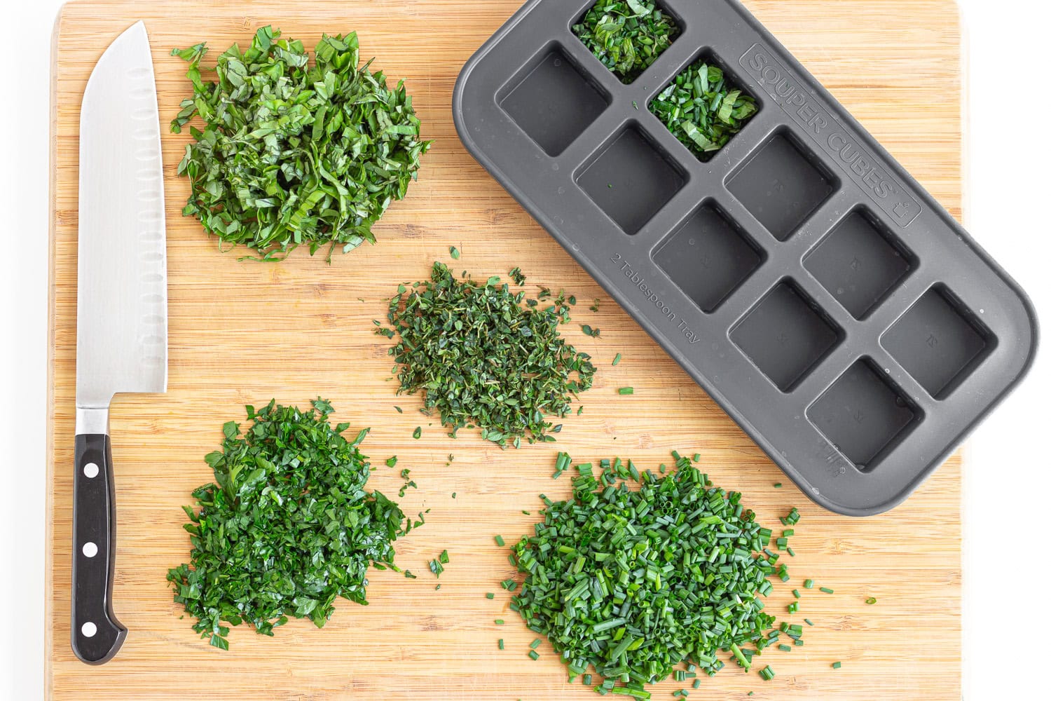 Cutting board with piles of chopped herbs, knife and silicone ice cube tray.
