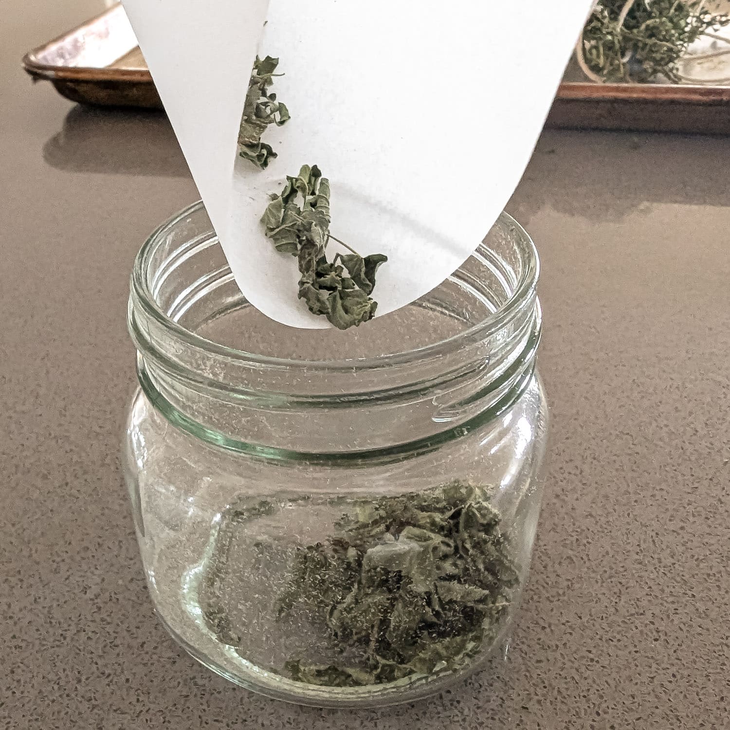 Dried herbs in parchment paper funnel being poured into a glass jar.