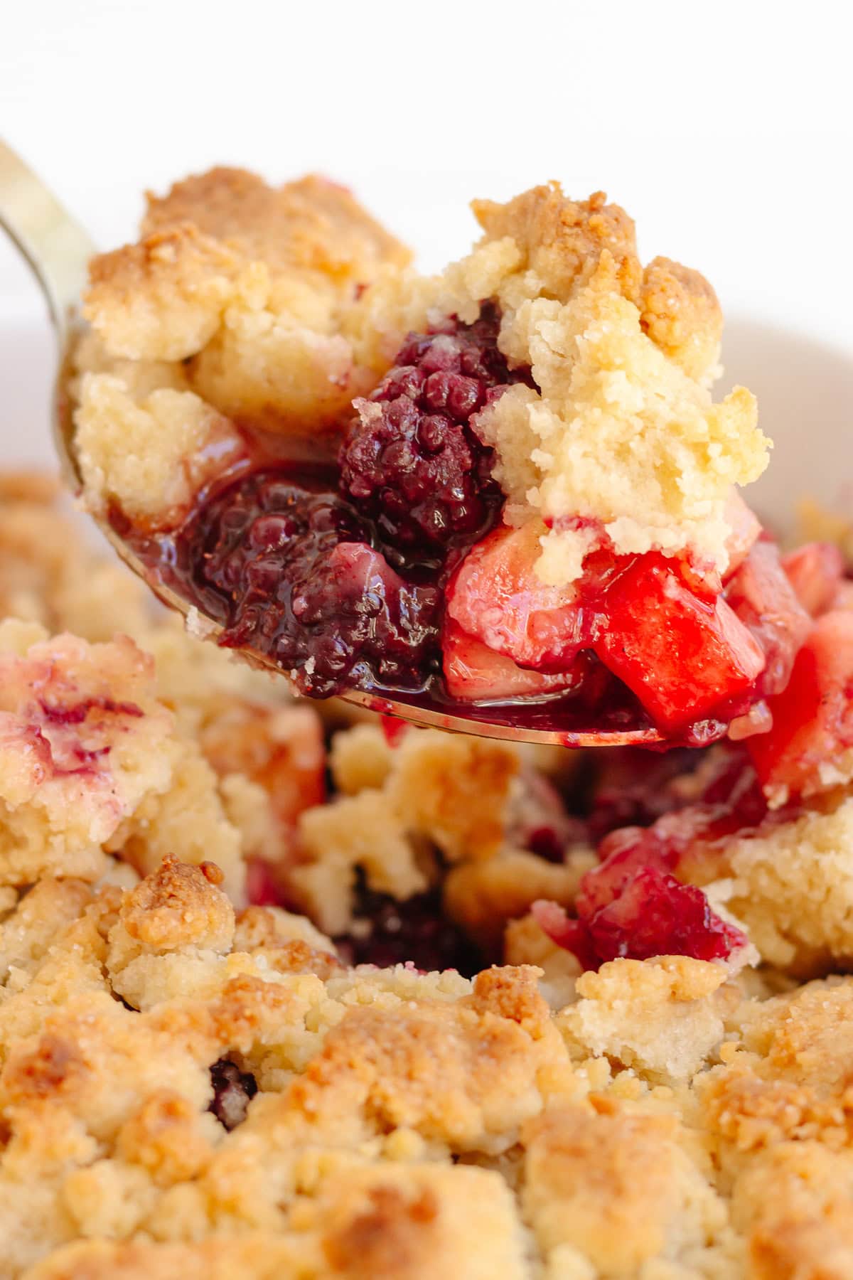 Scoop of apple and blackberry crumble being lifted from the baking dish.