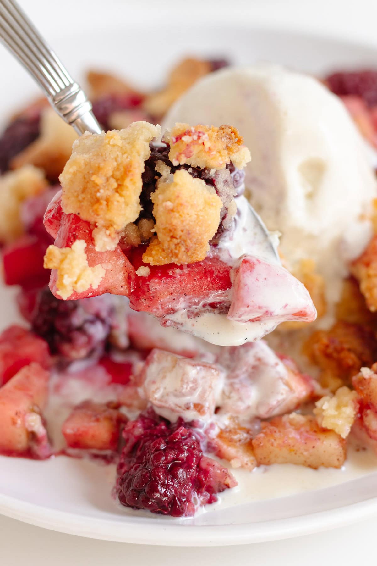 Spoonful of apple and blackberry crumble with ice cream being lifted from plate.