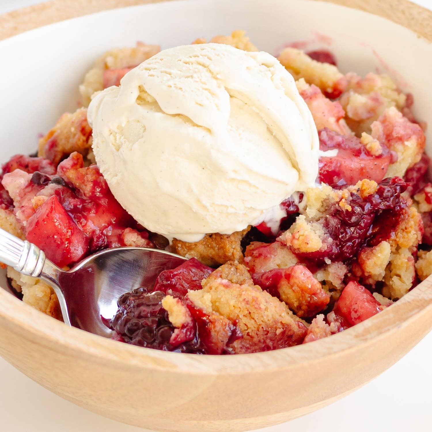 Apple and blackberry crumble in a wooden bowl topped with a scoop of vanilla ice cream.