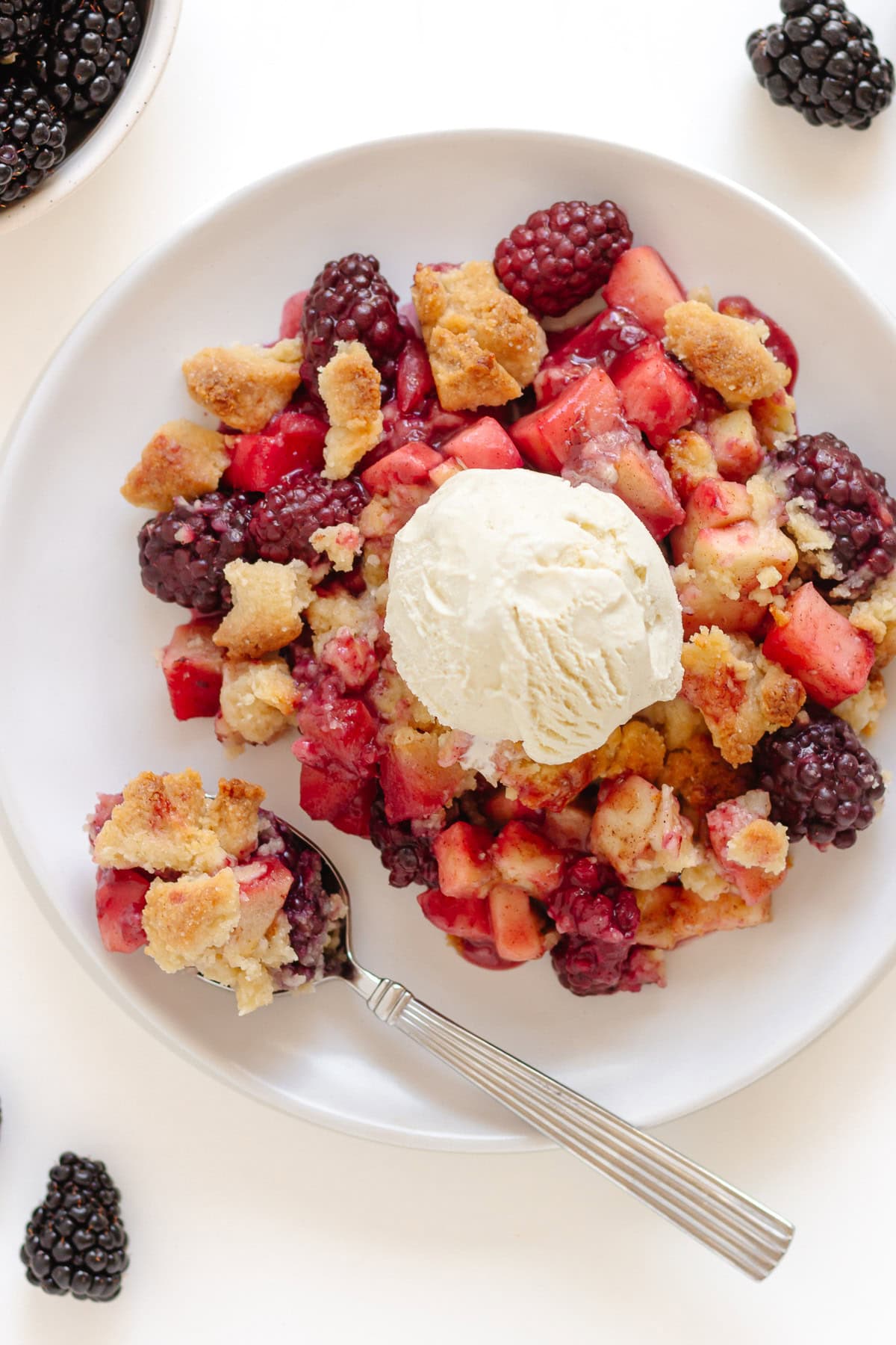 White plate of apple blackberry crumble topped with a scoop of vanilla ice cream and fresh blackberries scattered around.