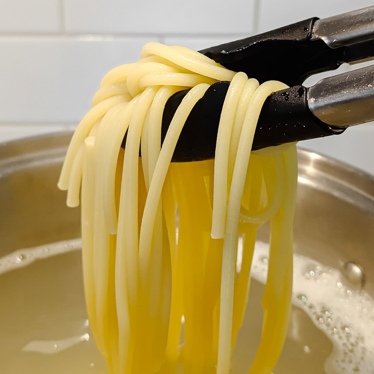 Tongs lifting cooked bucatini pasta out of a pot.