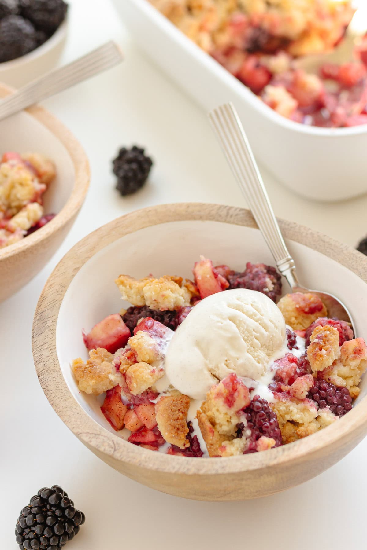 Homemade apple blackberry crumble in serving bowls topped with ice cream and baking dish in the background.