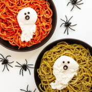 Two plates of spaghetti (one with tomato sauce and one with pesto) topped with burrata ghosts and surrounded by spiders.